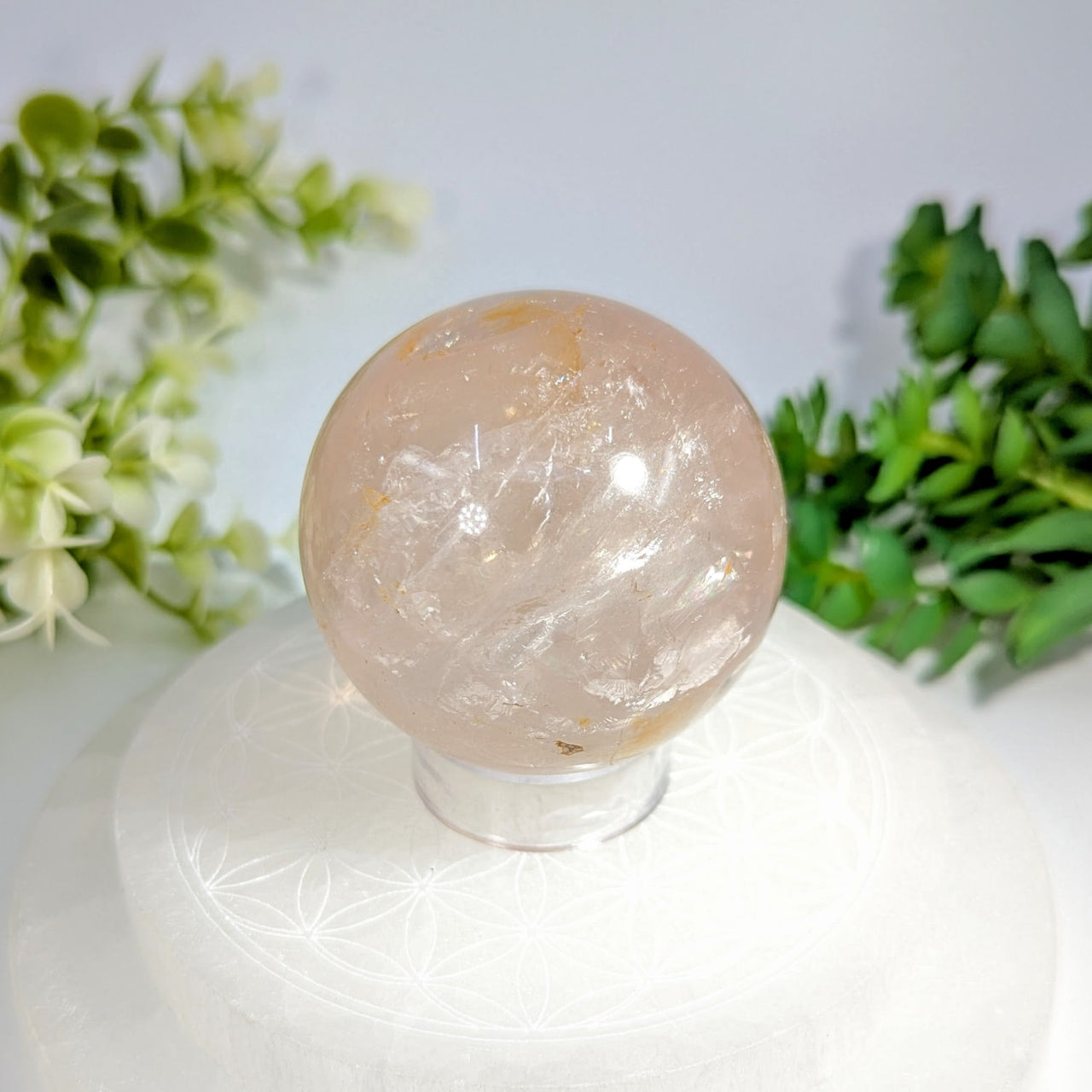 A translucent pink clear quartz sphere with hematoid golden inclusions displayed on a clear acrylic stand