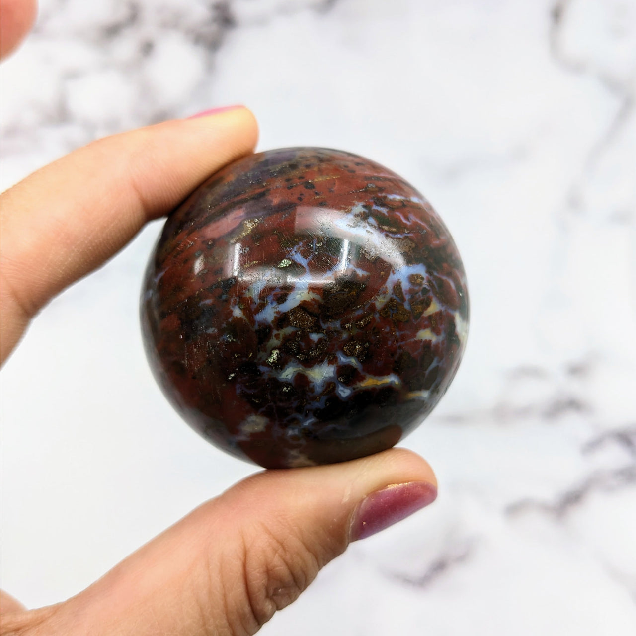 Hand holding Colorful Bloodstone Sphere #S062 on marble table