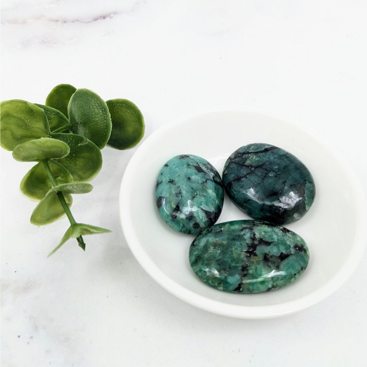 Three green palm stones in a white bowl next to a plant, part of Emerald 1 Collection