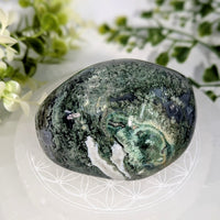 A polished green and black moss agate energy stone with white veining resting on a clear glass plate