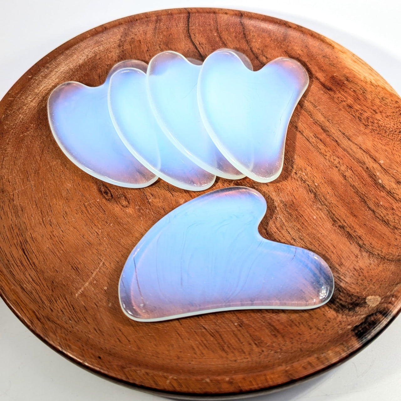 Five iridescent blue Opalite 3’’ gua sha massage tools on wooden plate