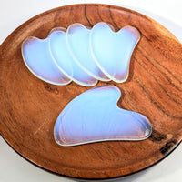 Five iridescent blue Opalite 3’’ gua sha massage tools on wooden plate