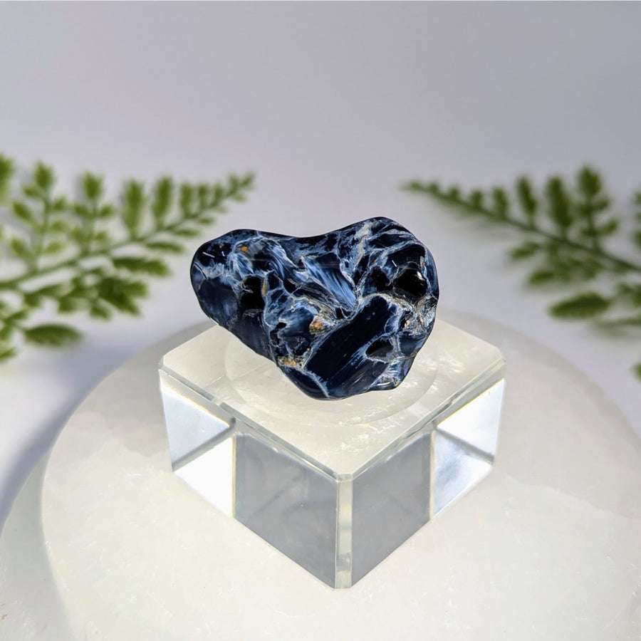 Pietersite 15 inch tumbled stone with polished dark blue and white marbled patterns displayed on clear acrylic pedestal for jewelry and crystals