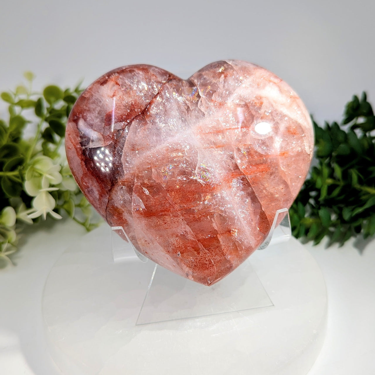 A polished 4 inch red hematoid quartz heart crystal with translucent pinkish red hue and internal patterns on acrylic stand