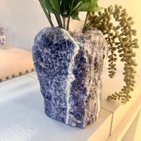 A rustic sodalite 8 inch blue and white rough stone vase holding green artificial foliage