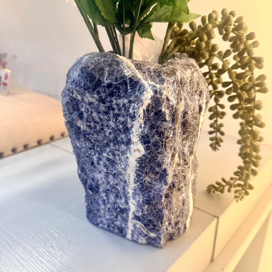 A rustic sodalite 8 inch blue and white rough stone vase holding green artificial foliage